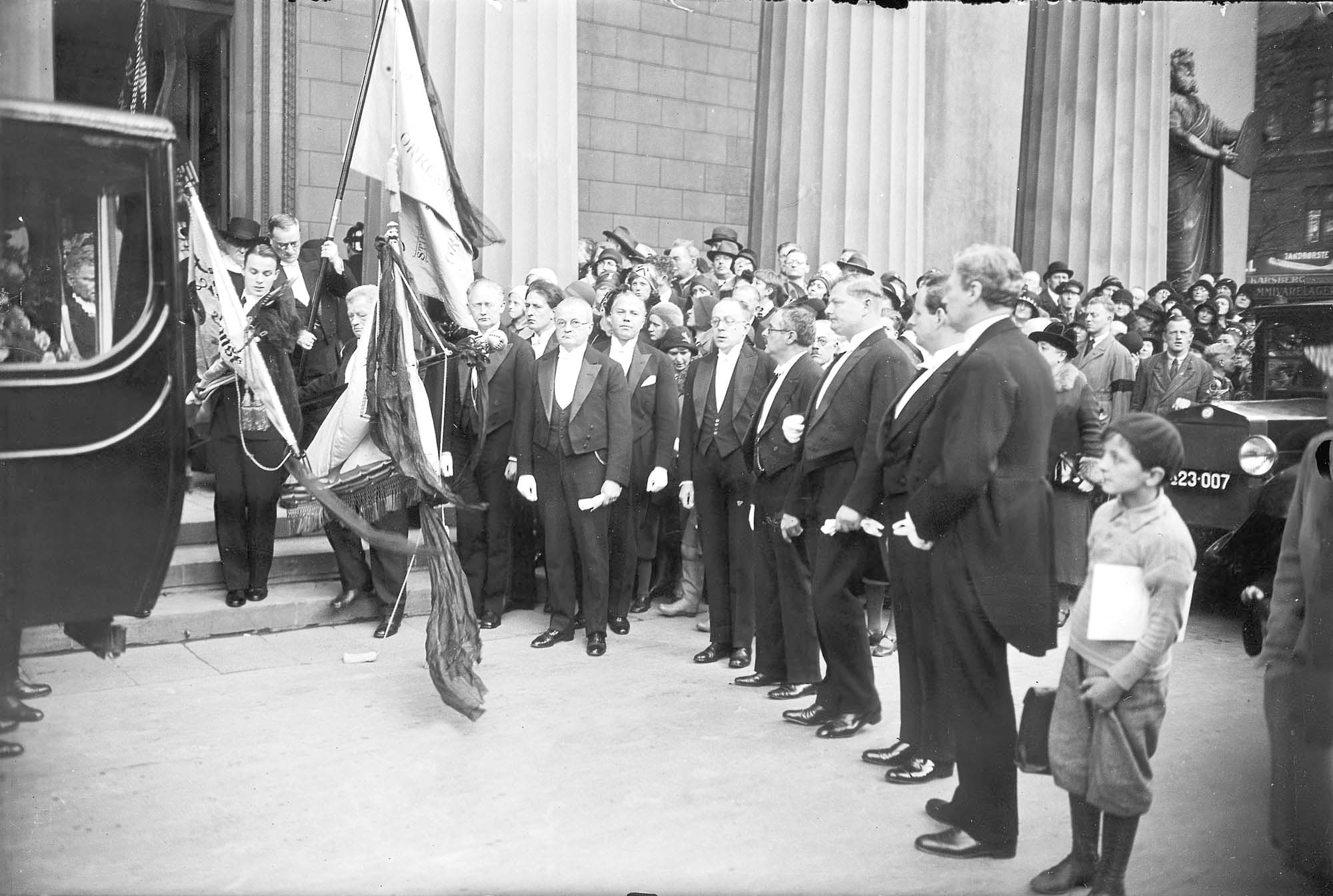 Blandt de kjoleklædte herrer ved rustvognen ses Peder Gram og Ebbe Hamerik (under den venstre søjle) samt Knud Jeppesen, Rudolph Simonsen og Poul Schierbeck.